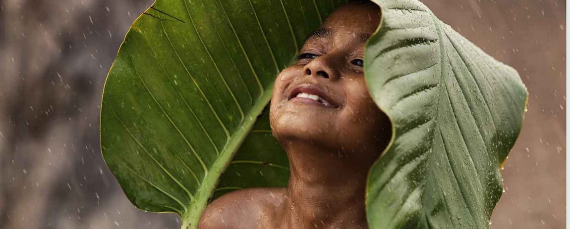 Un bambino si copre con un'ampia foglia dalla pioggia che lo bagna. Il suo viso, sereno, comunica tranquillità e fiducia.