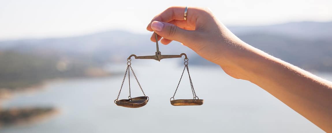 In primo piano una mano sorregge un bilancino meccanico dove, uno dei due piatti è un po' ammaccato, come se sottolineassi la precarietà dell'equilibrio.