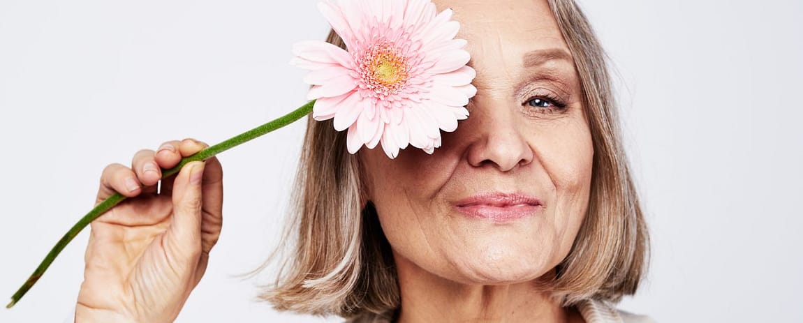 Donna matura sorridente con un fiore rosa davanti a un occhio, immagine dedicata alla menopausa e al benessere psicologico.