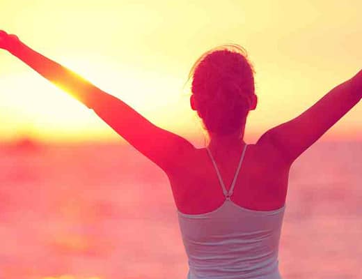 Una donna con le braccia al cielo guarda il sole che sorge.