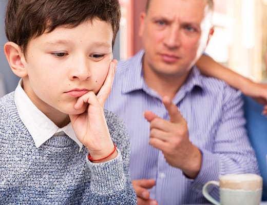 Un bimbo imbonciato in primo piano e una coppia genitoriale sullo sfondo. Il papà con un atteggiamento rimproverante.