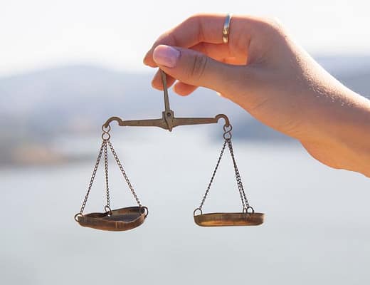 In primo piano una mano sorregge un bilancino meccanico dove, uno dei due piatti è un po' ammaccato, come se sottolineassi la precarietà dell'equilibrio.