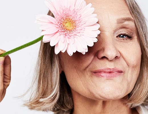 Donna matura sorridente con un fiore rosa davanti a un occhio, immagine dedicata alla menopausa e al benessere psicologico.